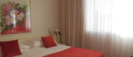 Habitación de hotel con cama, almohadas rojas, cabecera blanca, un cuadro de flores, una lámpara de noche y una ventana con cortinas traslúcidas.