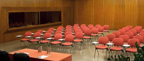 Un salón de conferencias con filas de sillas rojas y mesas, paneles de madera en las paredes y una pantalla grande al frente.