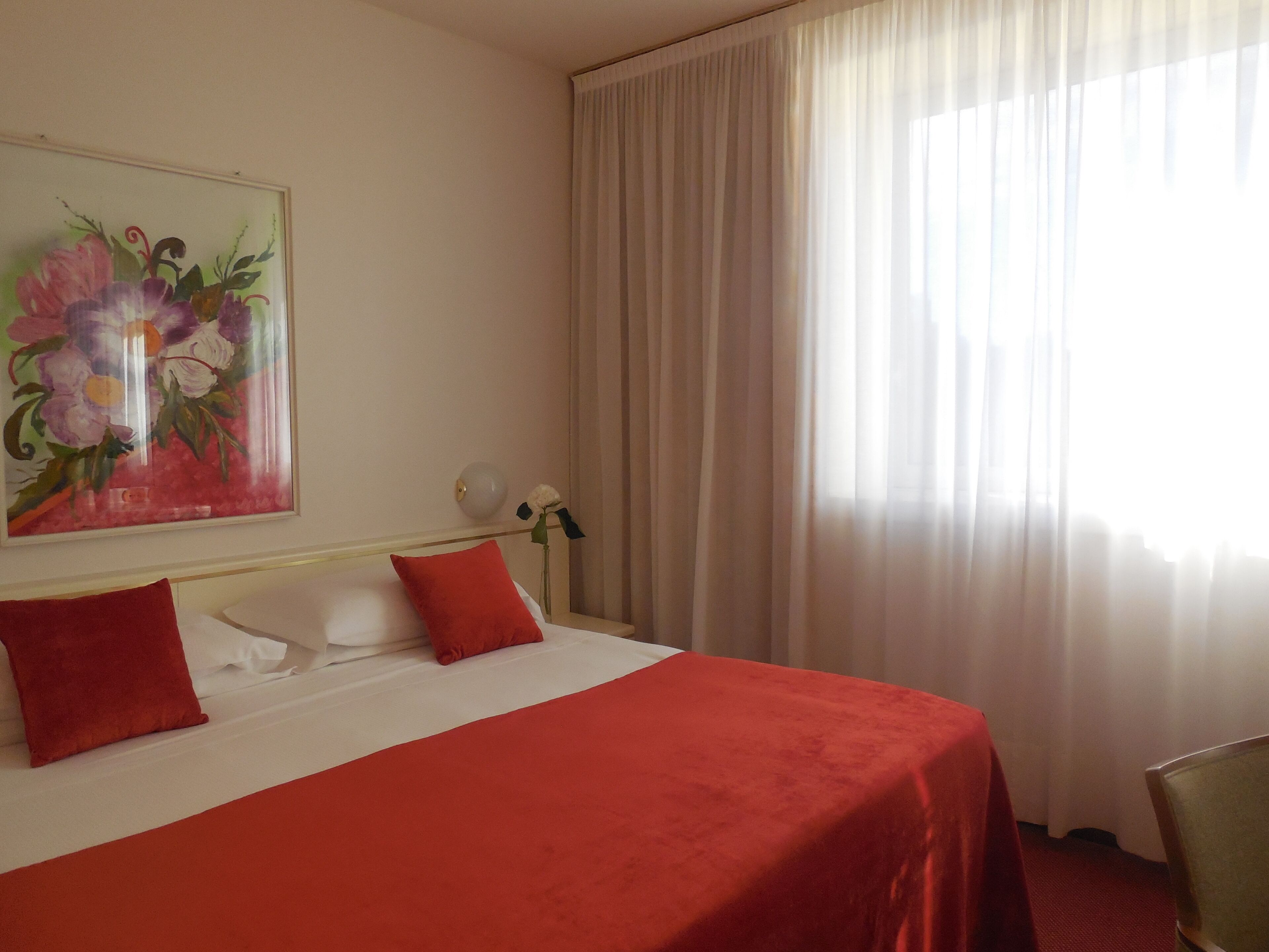 Ein Hotelzimmer mit einem Bett, einem Blumenbild, einer Vase mit einer Blume und einem Fenster mit Vorhängen.