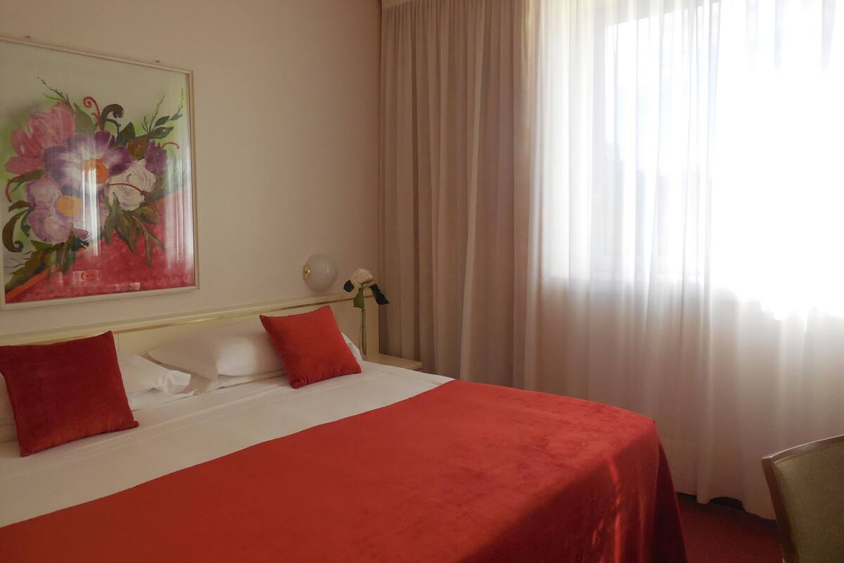 A hotel room with a bed, red pillows, a white headboard, a painting of flowers, a vase with a flower, and a window with sheer curtains.