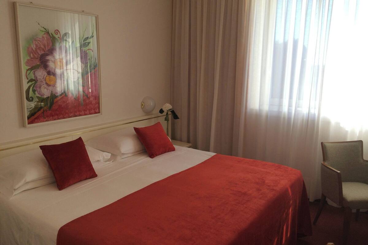 A hotel room with a large bed, red pillows, a white headboard, a painting of flowers on the wall, a nightstand with a lamp, and a chair.