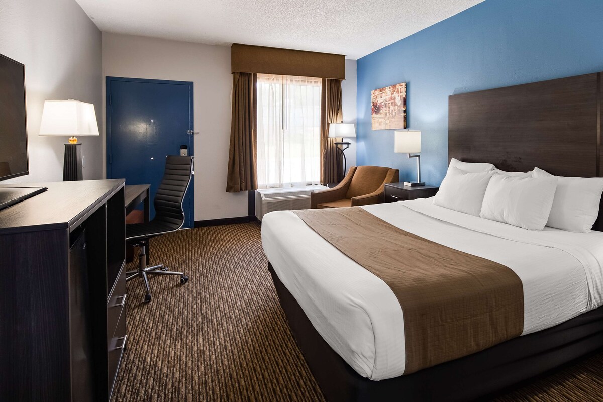 A hotel room with a large bed, a desk with a computer, a chair, and a window with curtains.