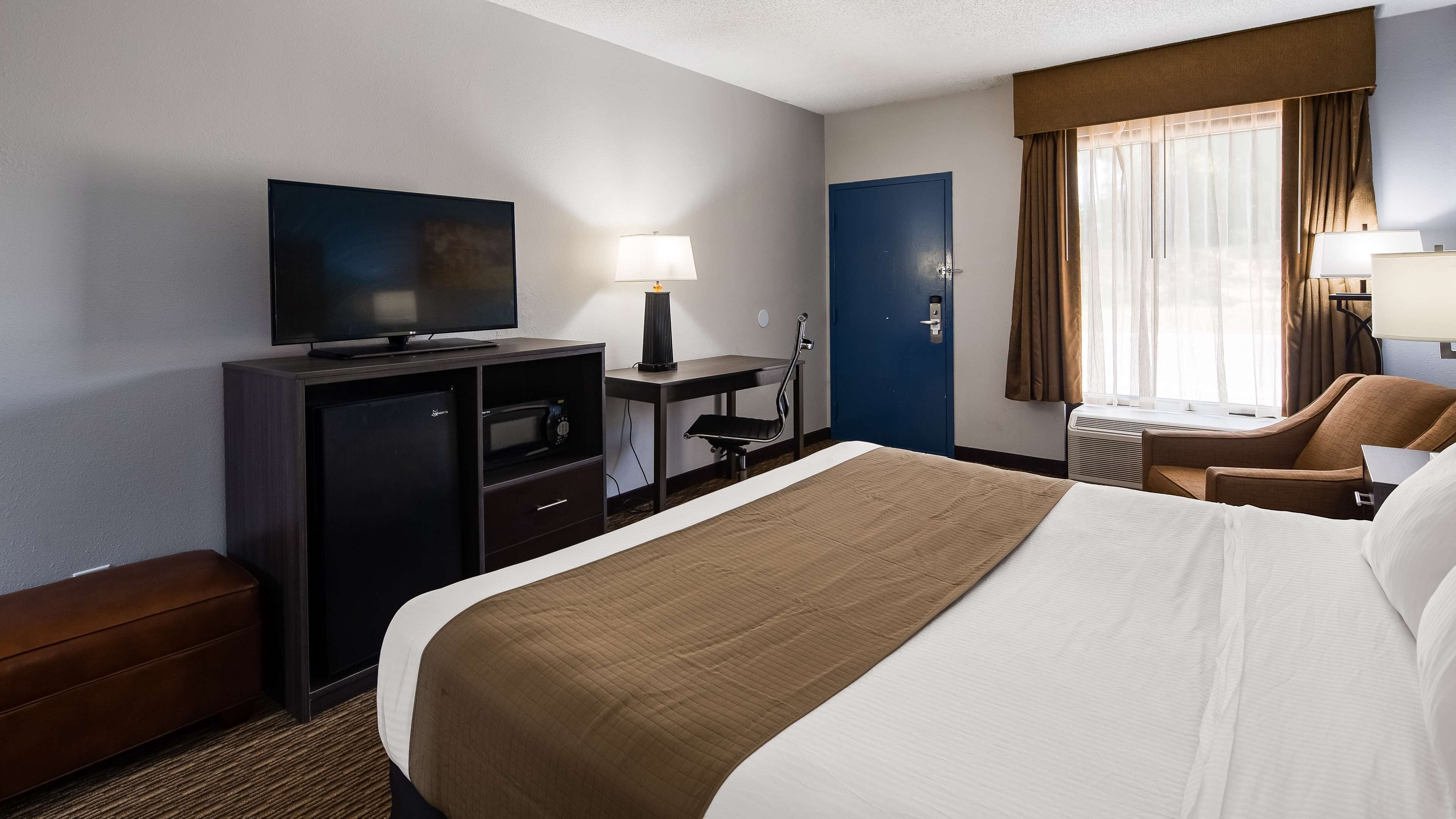Habitación de hotel con cama, escritorio con computadora, televisión, minibar y ventana con cortinas.