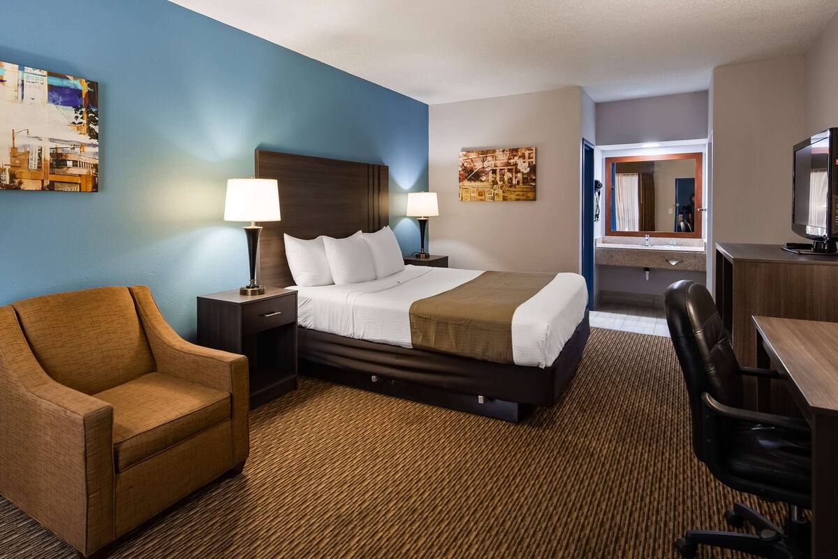 A hotel room with a bed, two bedside tables with lamps, a brown armchair, a desk with a chair, and artwork on the walls.