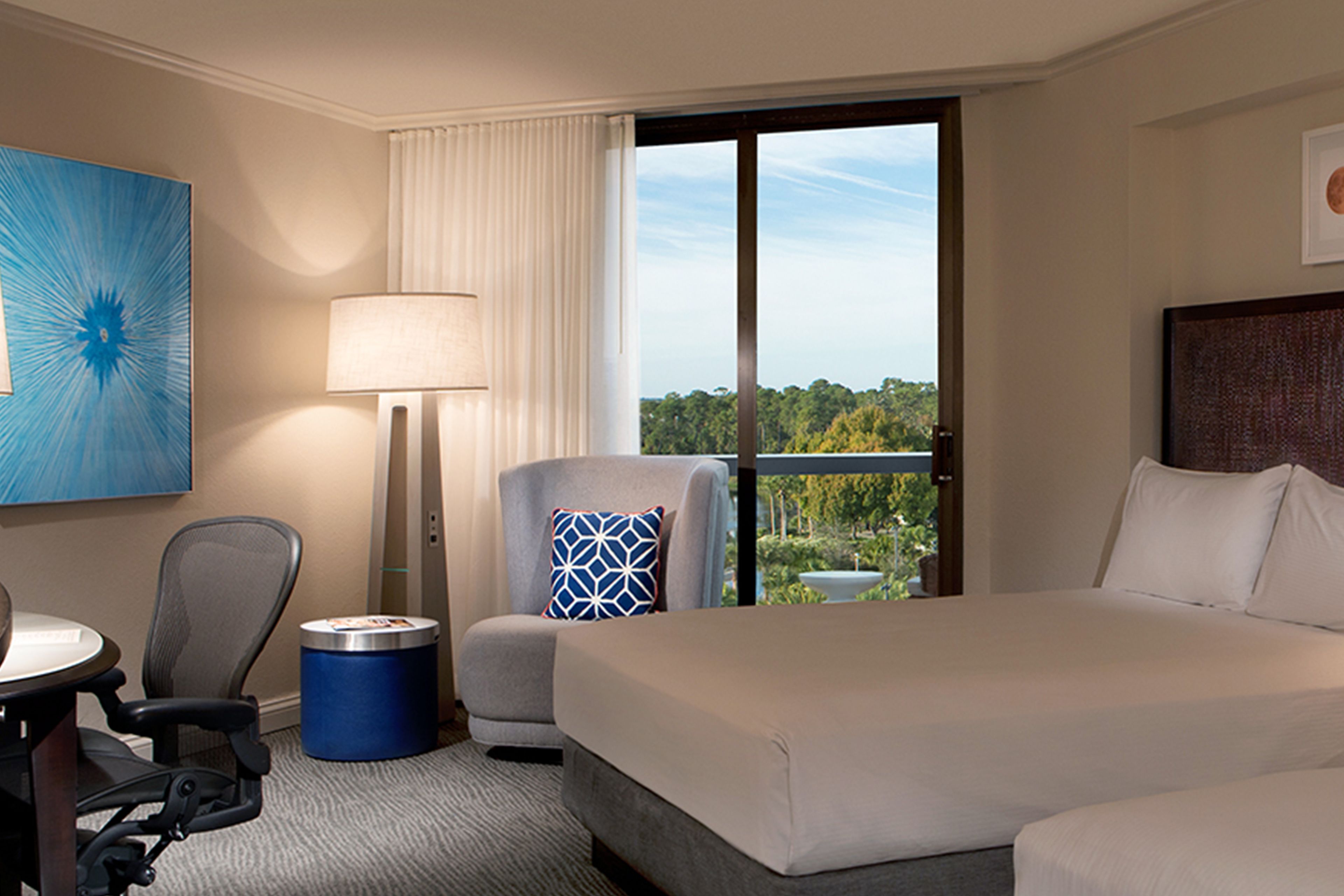 A hotel room with a large bed, a desk with a chair, a lamp, and a view of greenery through the window.