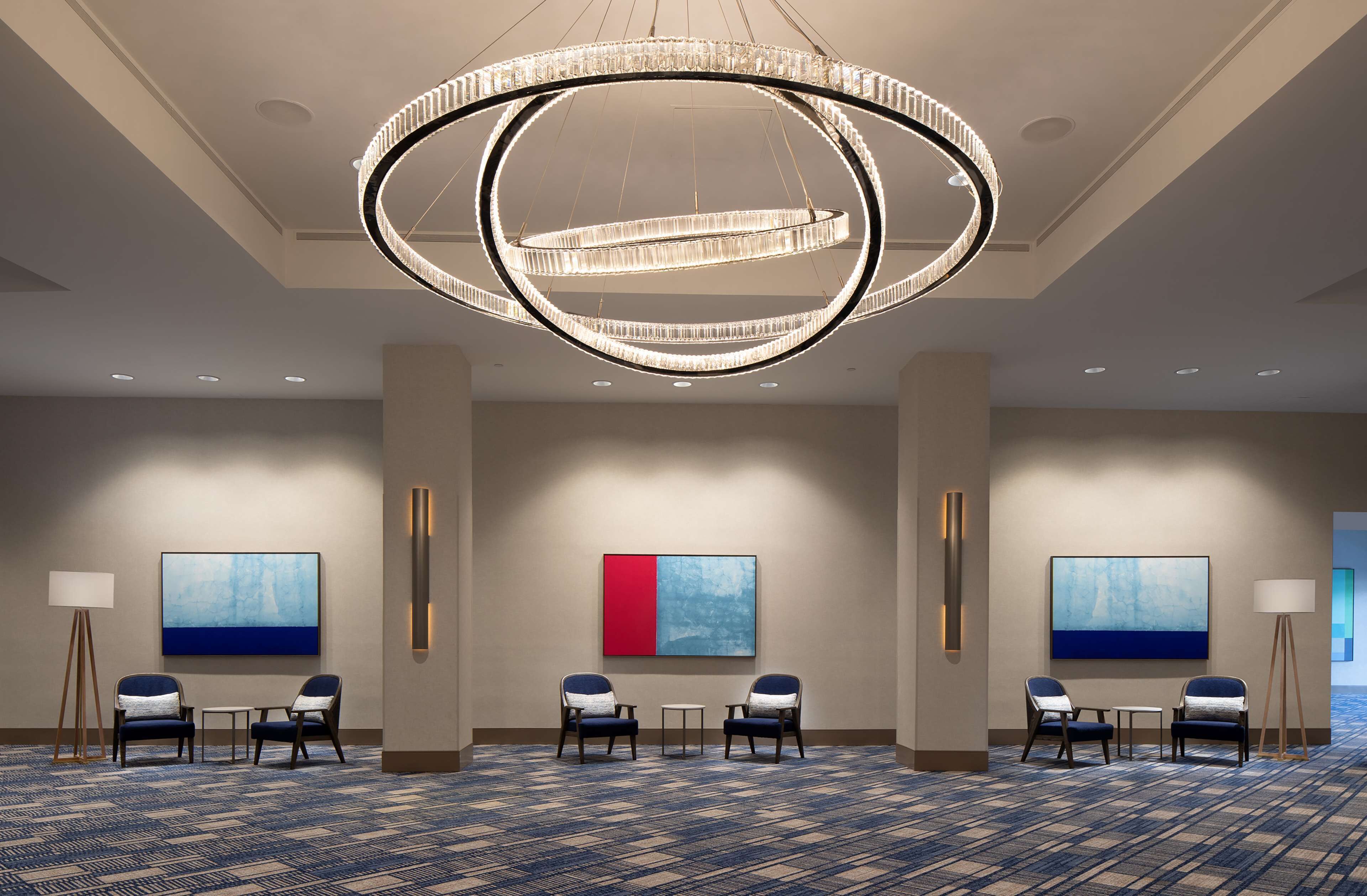 A hotel lobby with a large circular chandelier, two paintings on the walls, and a carpeted floor.