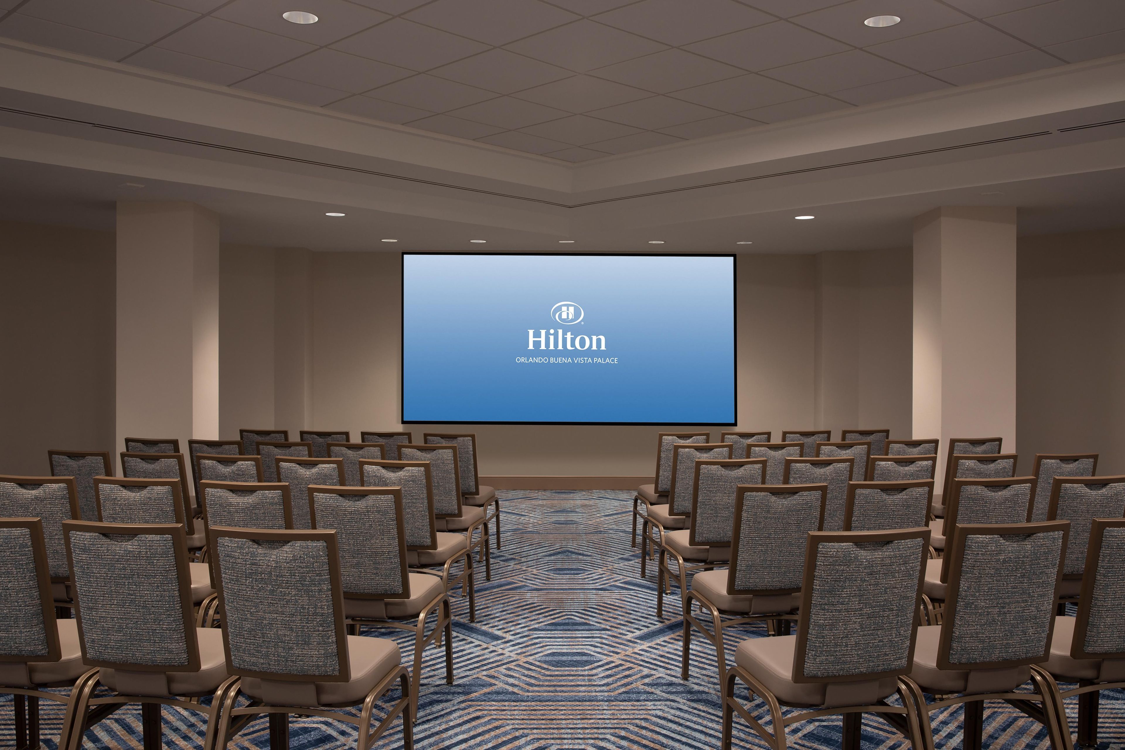 Un salón de conferencias con filas de sillas mirando hacia una pantalla que muestra el logotipo de Hilton.