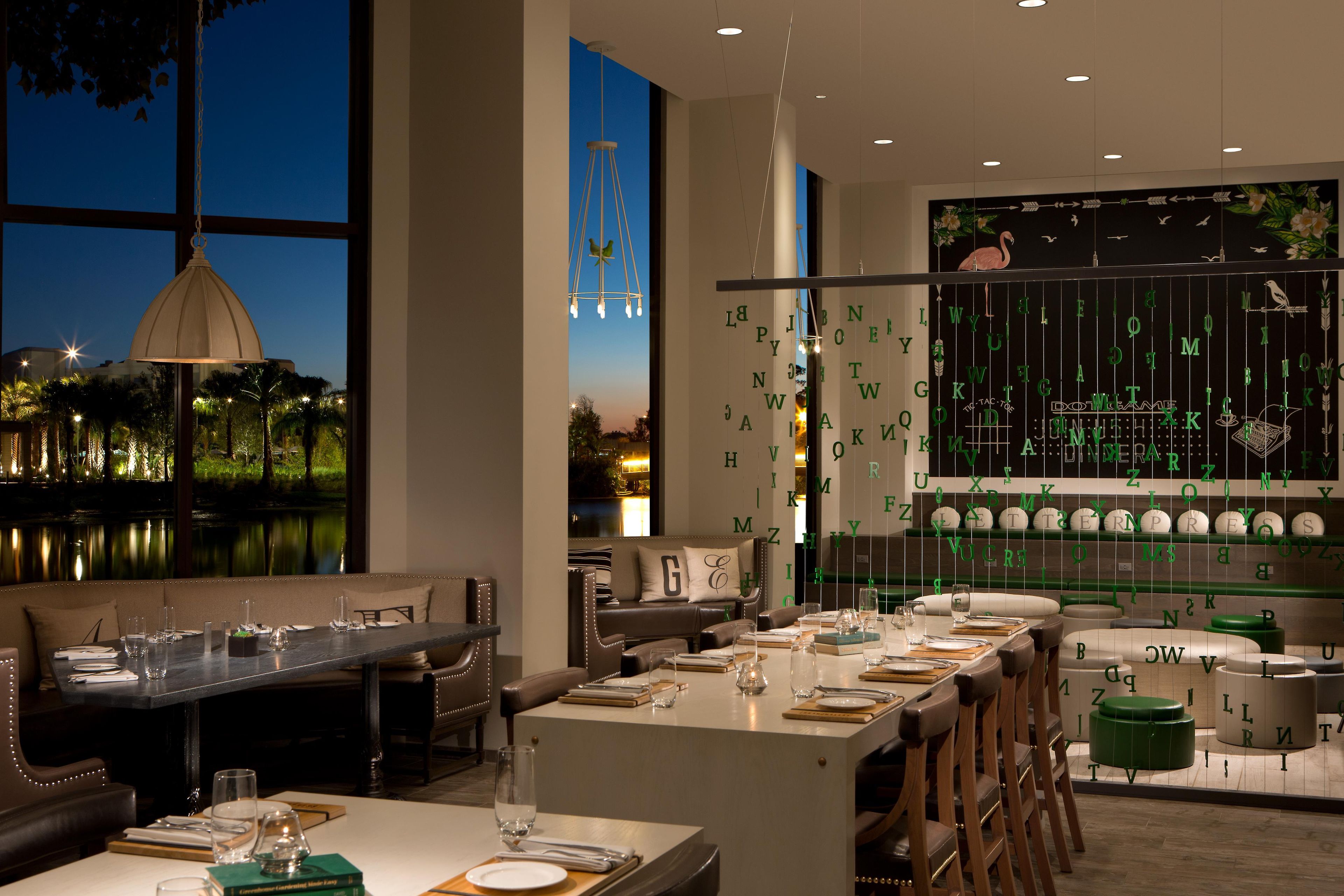 A restaurant with a view of a body of water, a dining area with tables set for two, and a wall with hanging letters.