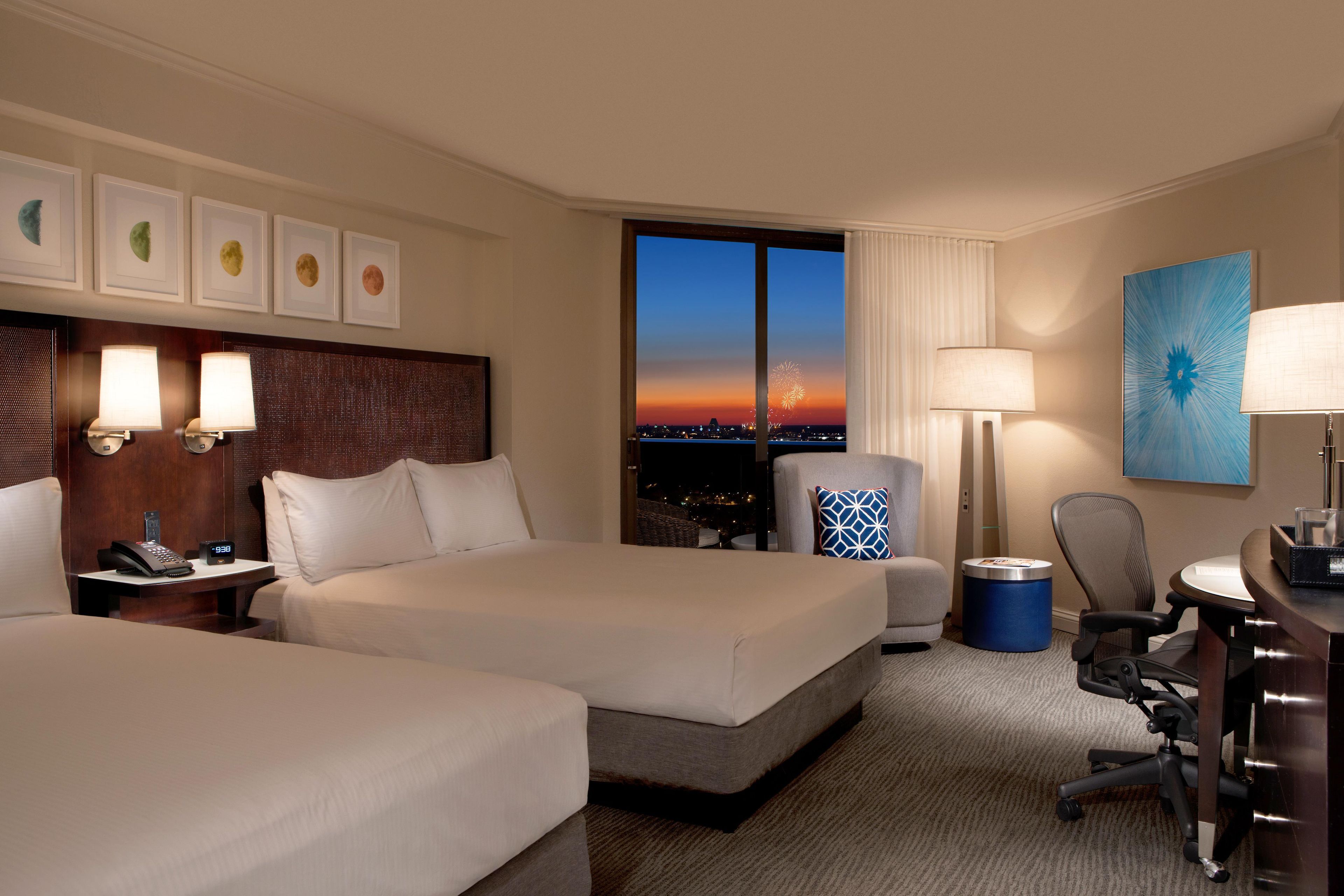 A hotel room with two beds, a desk, a chair, a lamp, a phone, and a view of the city at sunset.