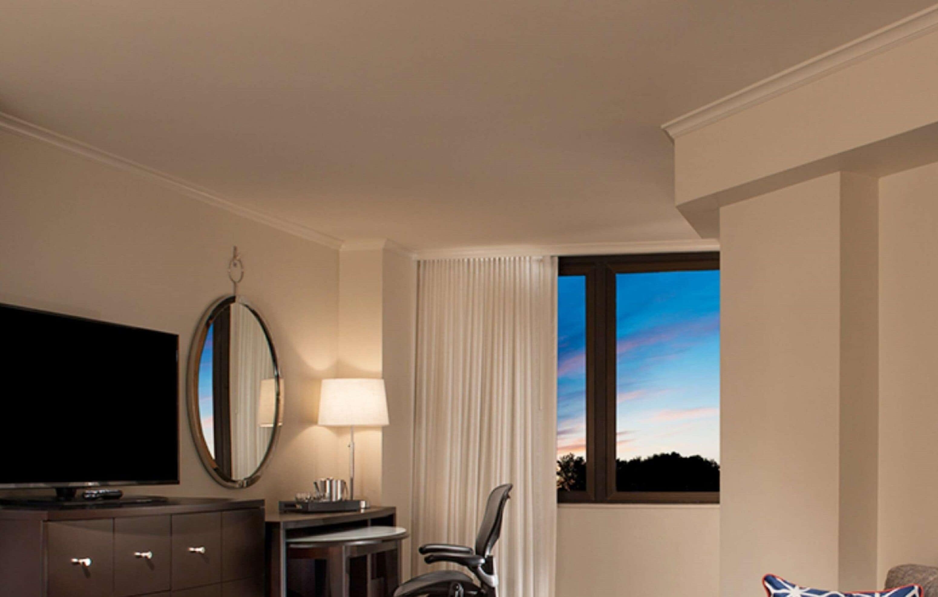 A hotel room with a large flat-screen TV, a desk with a lamp, a chair, and a window with a view of the sky.