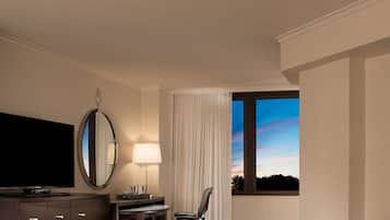 A hotel room with a large flat-screen TV, a desk with a lamp, a chair, and a window with a view of the sky.