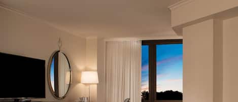 A hotel room with a large flat-screen TV, a desk with a lamp, a chair, and a window with a view of the sky.