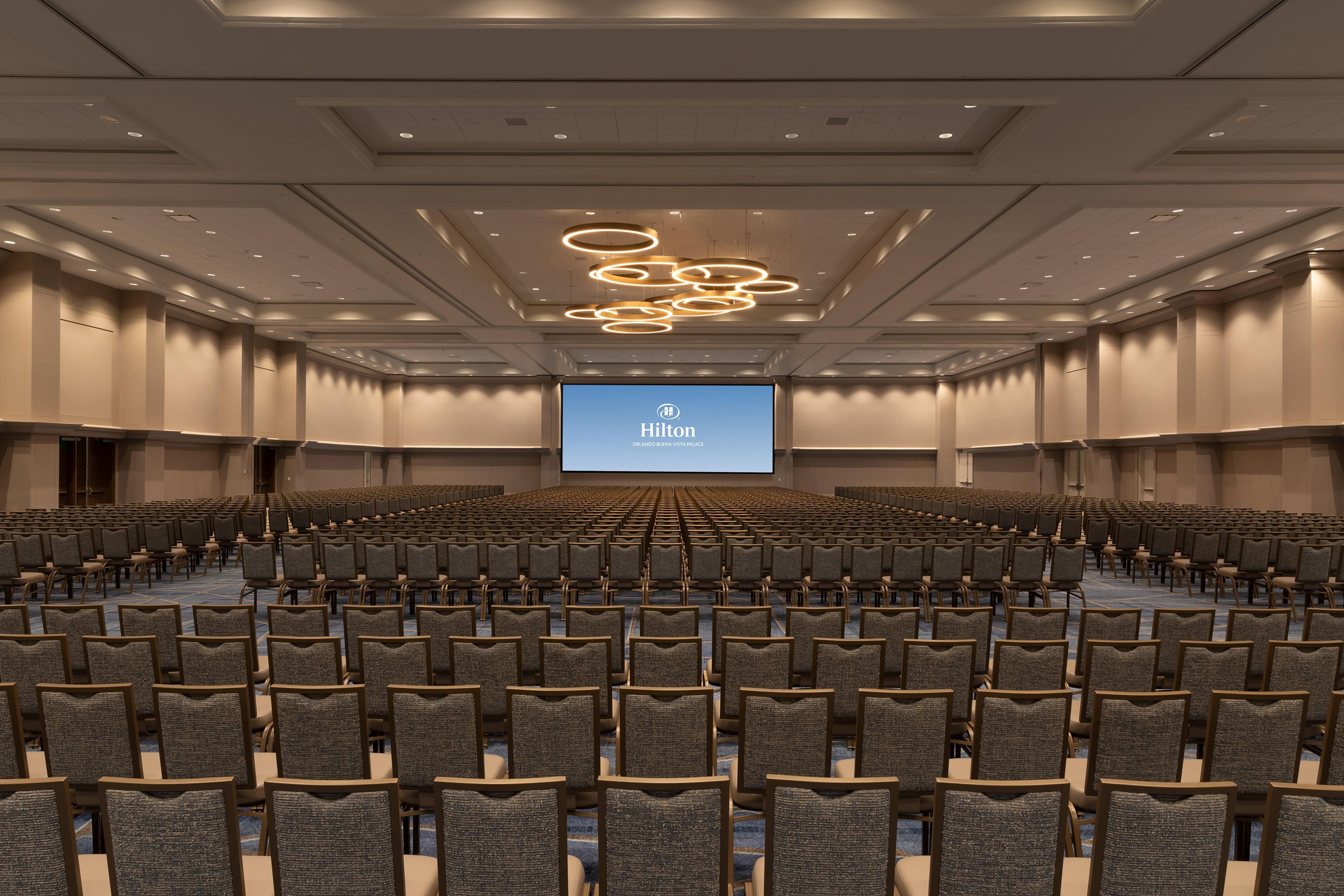 A large conference room with rows of chairs, a stage with a screen displaying the Hilton logo, and modern lighting fixtures.
