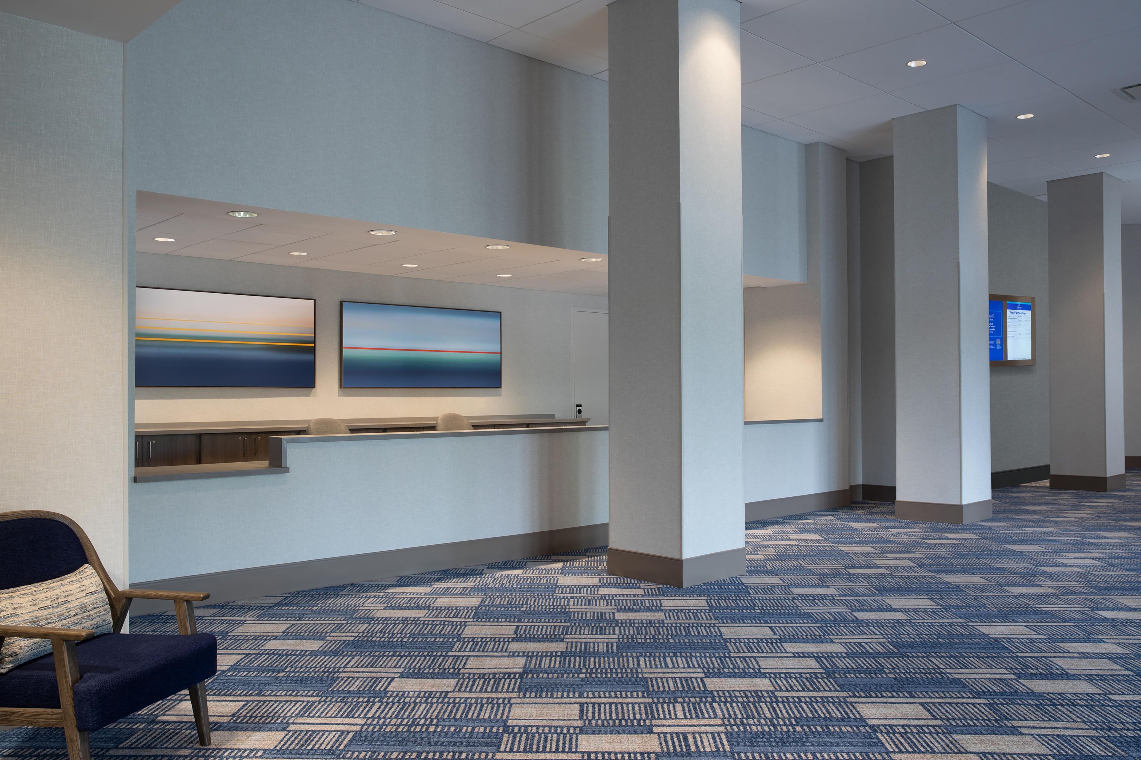 Un espace d’accueil d’hôtel comprenant un comptoir de réception, deux œuvres d’art encadrées, une moquette bleue à motifs et une chaise en bois.