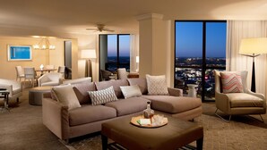 A modern hotel room with a large sofa, a coffee table with a tray, a side table, a lamp, and a view of the city at dusk.