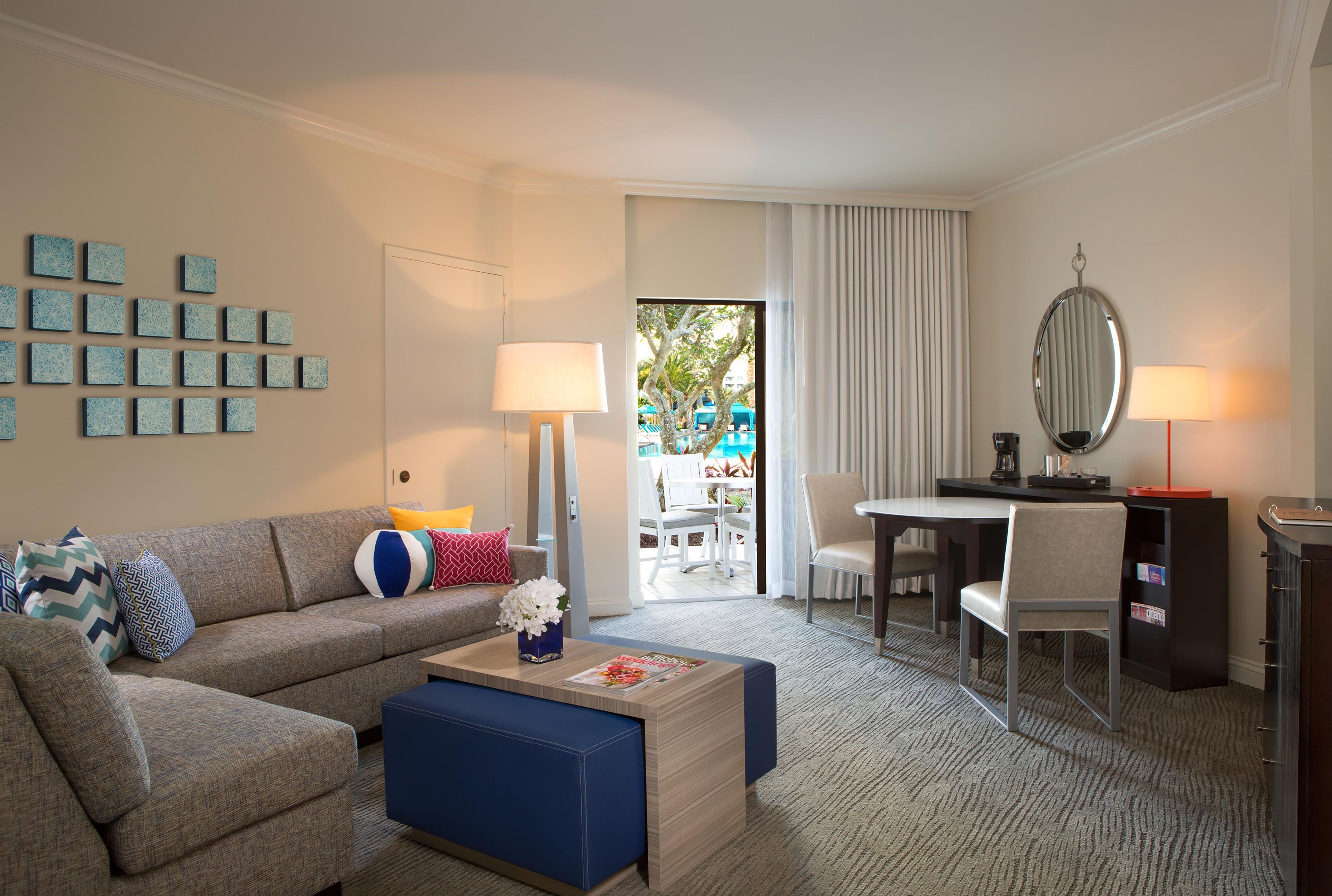 A hotel room with a sofa, a coffee table, a desk, and a chair. There is a view of an outdoor area with a pool.