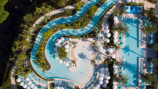 En vy från ovan av ett semesterområde med en slingrande pool, solstolar och palmer.