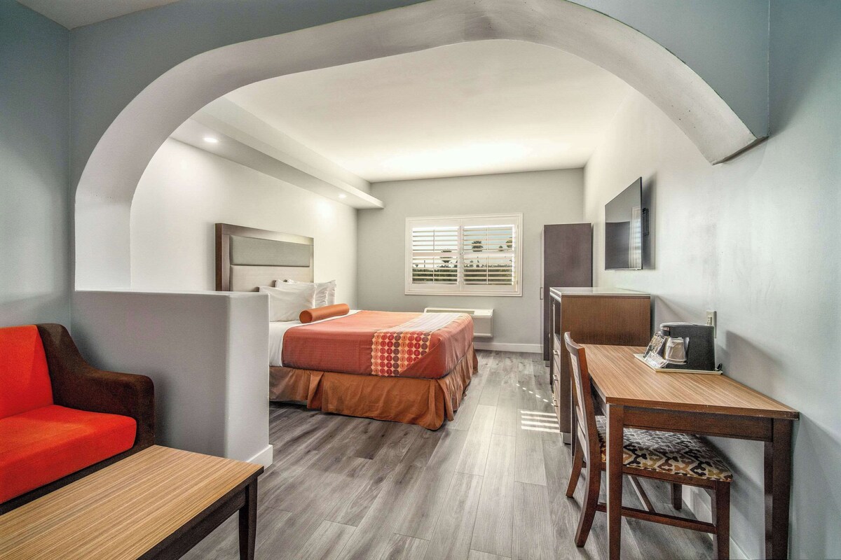 A compact hotel room with a bed, a desk, a chair, a sofa, and a TV mounted on the wall.