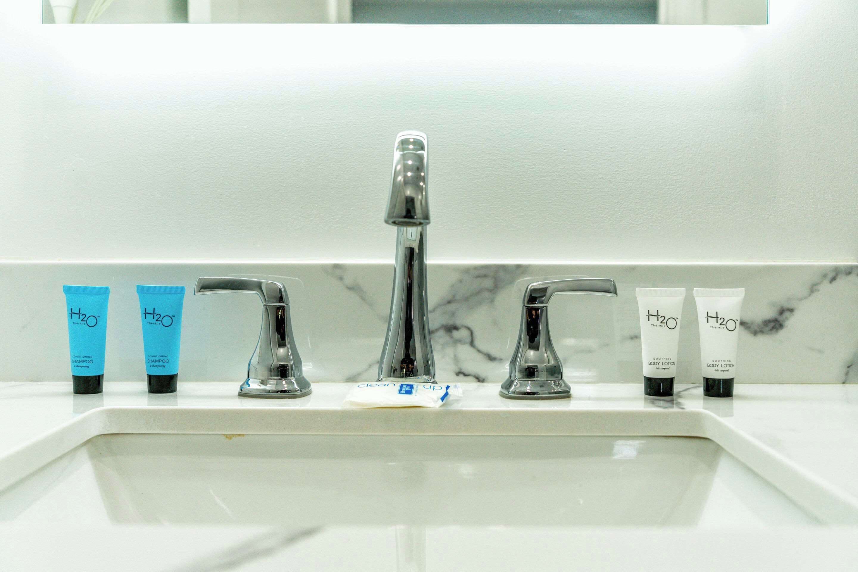 Un lavamanos con dos tubos de loción corporal H2O azul y blanco, una botella de vidrio transparente y un dispensador de jabón blanco.