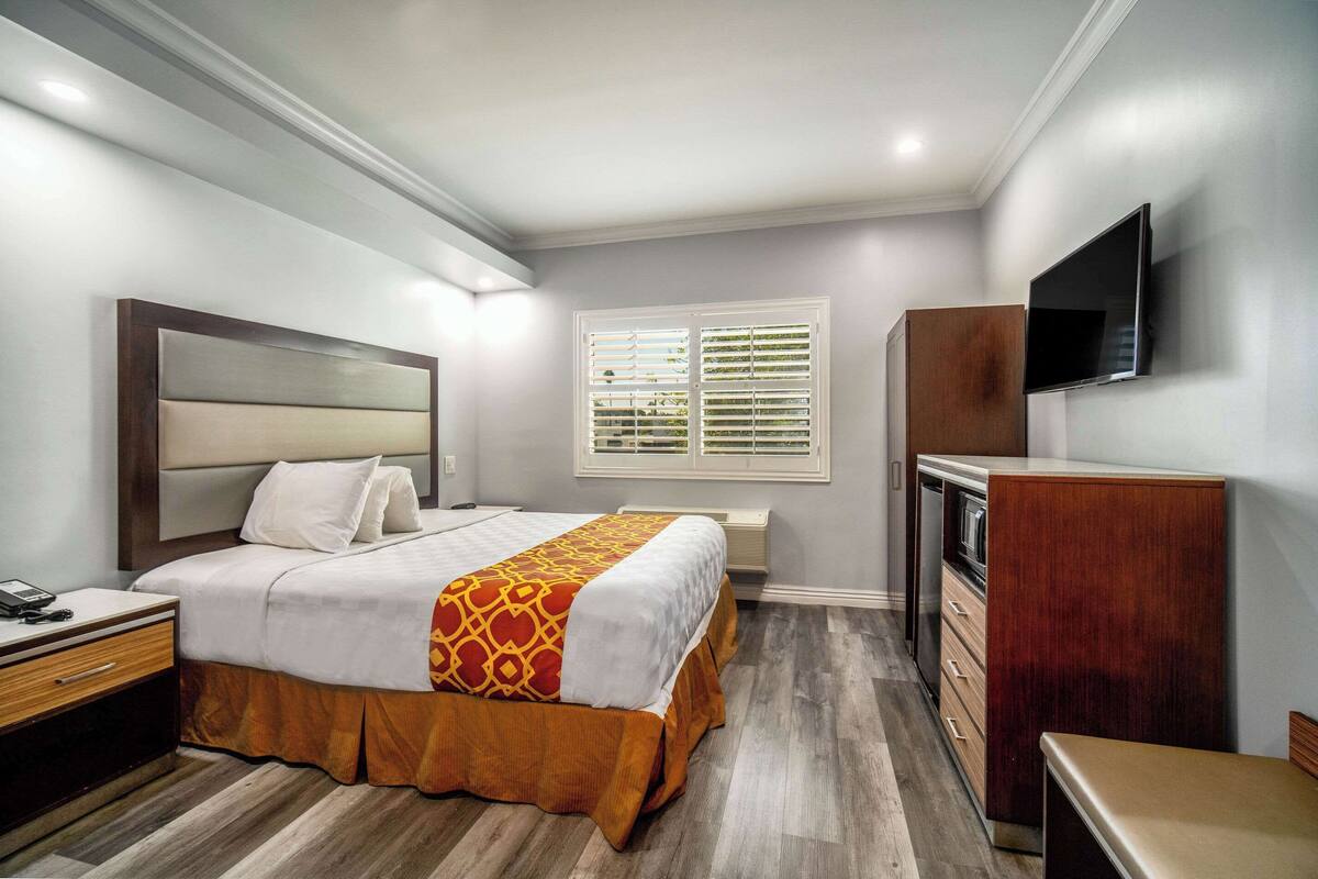 Een hotelkamer met een bed, nachtkastje, kaptafel, televisie en raam met zonwering.