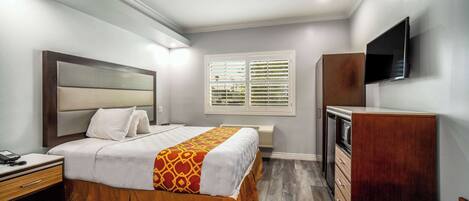 Habitación de hotel con cama, mesita de noche, cómoda, televisor y ventana con persianas.