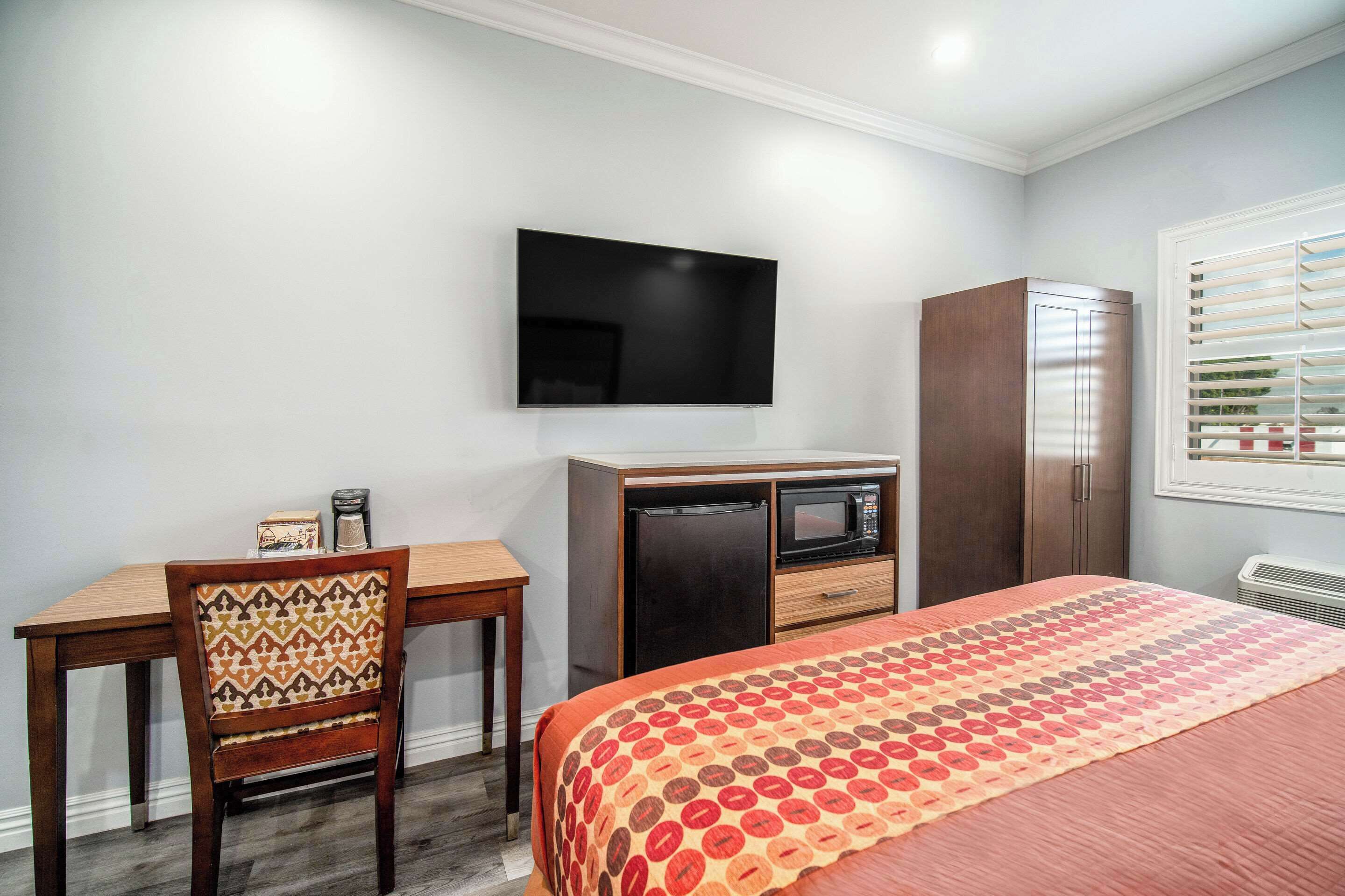 A compact hotel room with a bed, a desk with a chair, a TV mounted on the wall, a wardrobe, and a window with blinds.