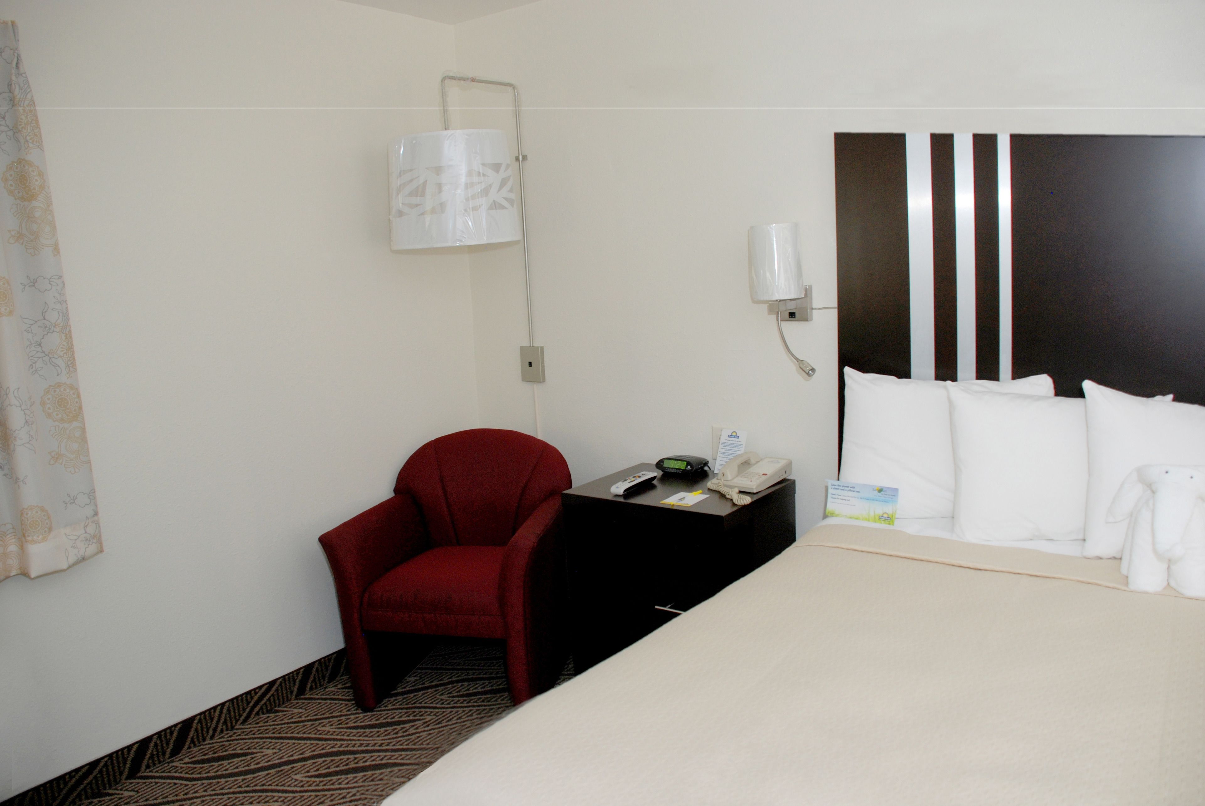 Una habitación de hotel con una cama, una silla roja, una mesita con un teléfono y una lámpara, y una lámpara de pared.