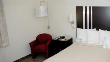 Una habitación de hotel con una cama, una silla roja, una mesita con un teléfono y una lámpara, y una lámpara de pared.
