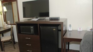 A hotel room with a desk, a television, a microwave, a refrigerator, a chair, and a trash bin.