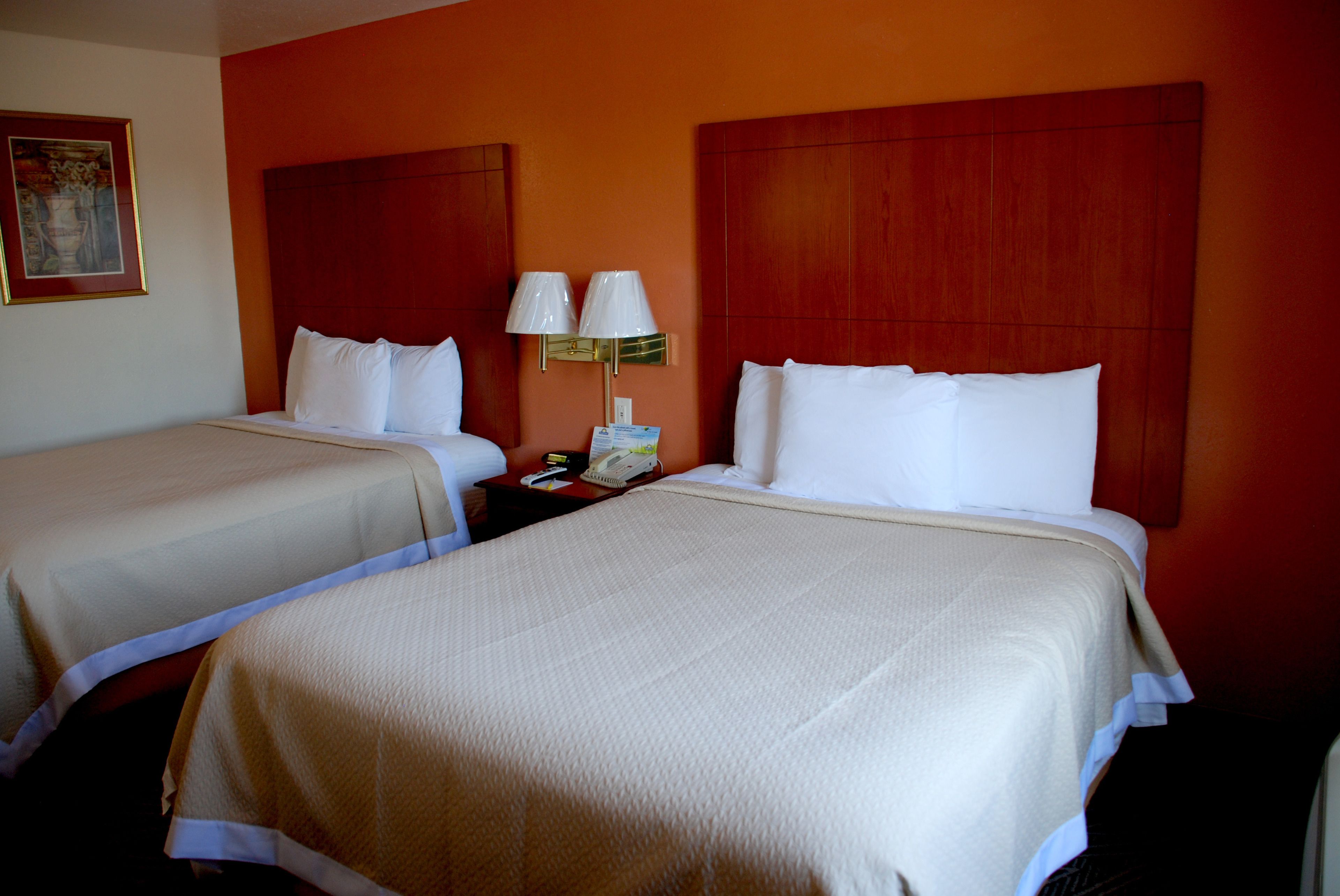 A hotel room with two beds, each with white linens and a wooden headboard. There is a nightstand with a lamp between the beds.