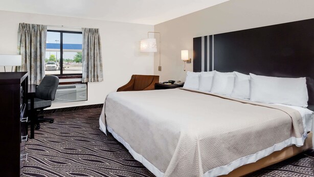 A hotel room with a large bed, a desk with a chair, a window with curtains, and a view of a parking lot.