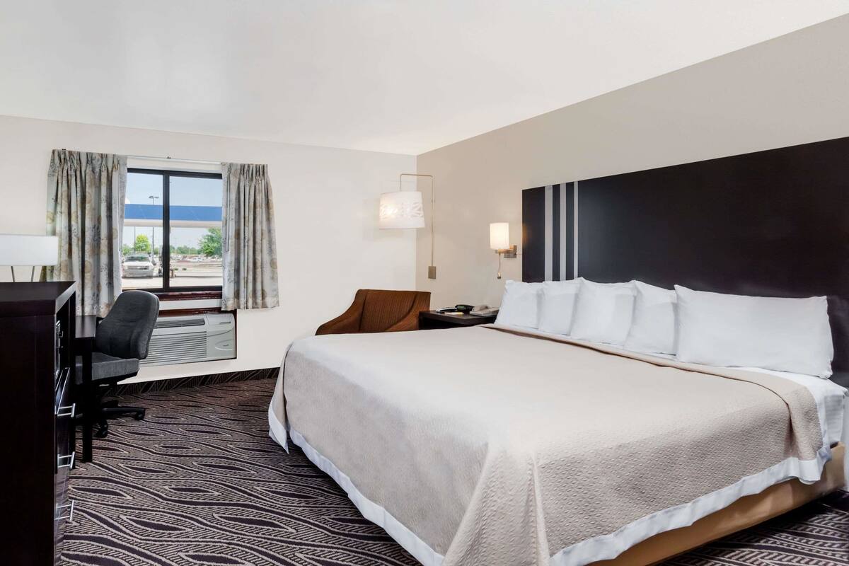 A hotel room with a large bed, a desk with a chair, a window with curtains, and a view of a parking lot.