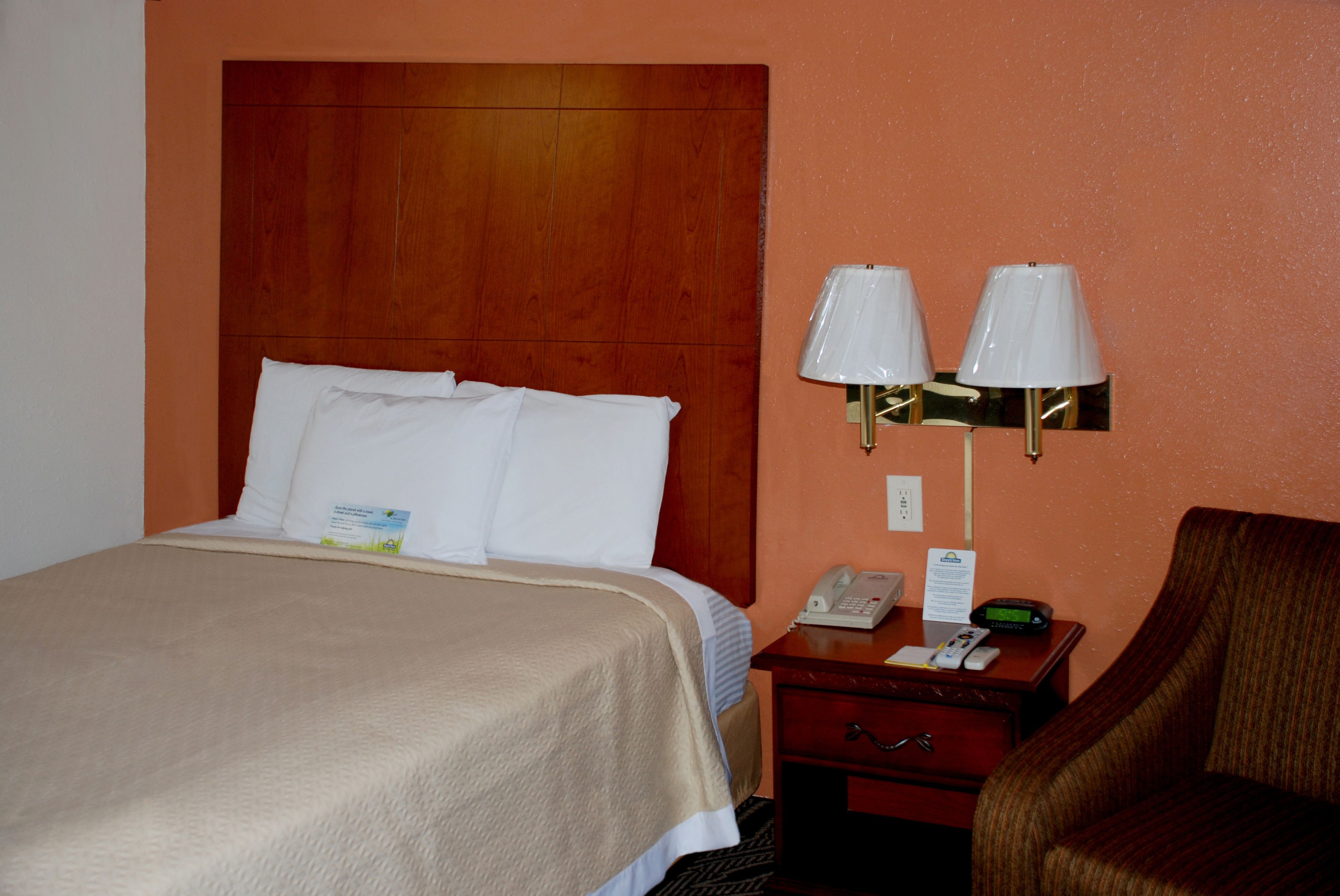Una habitación de hotel con cabecera de madera, cama con ropa de cama blanca, mesita de noche con lámpara, teléfono y una silla.