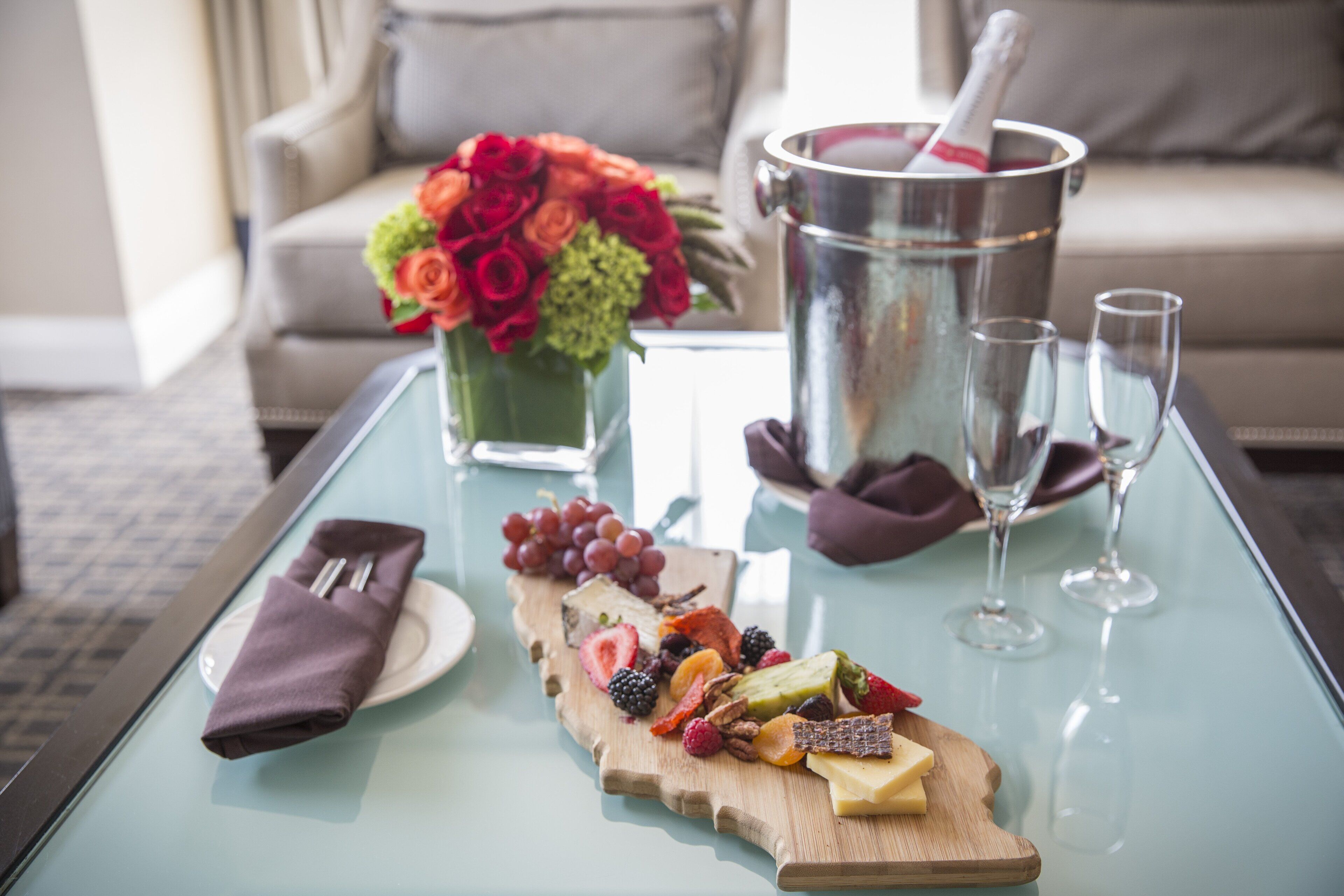 Una mesa con superficie de vidrio que exhibe una selección de quesos, frutas y una botella de champán. También se observa un florero con flores y un cubo para champán.