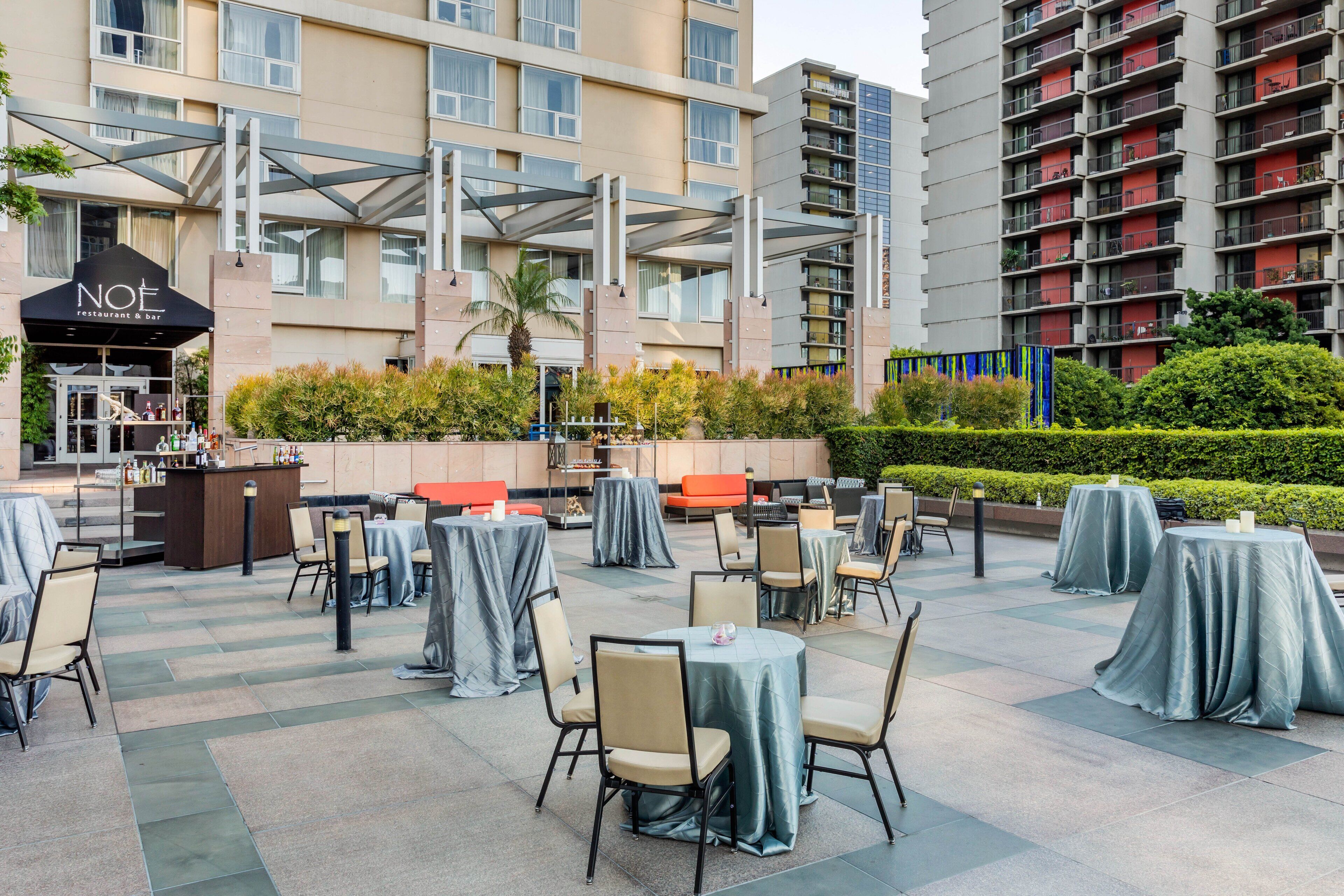 Un espace de restauration en plein air avec des tables et des chaises, un bar, et un bâtiment à plusieurs étages en arrière-plan.