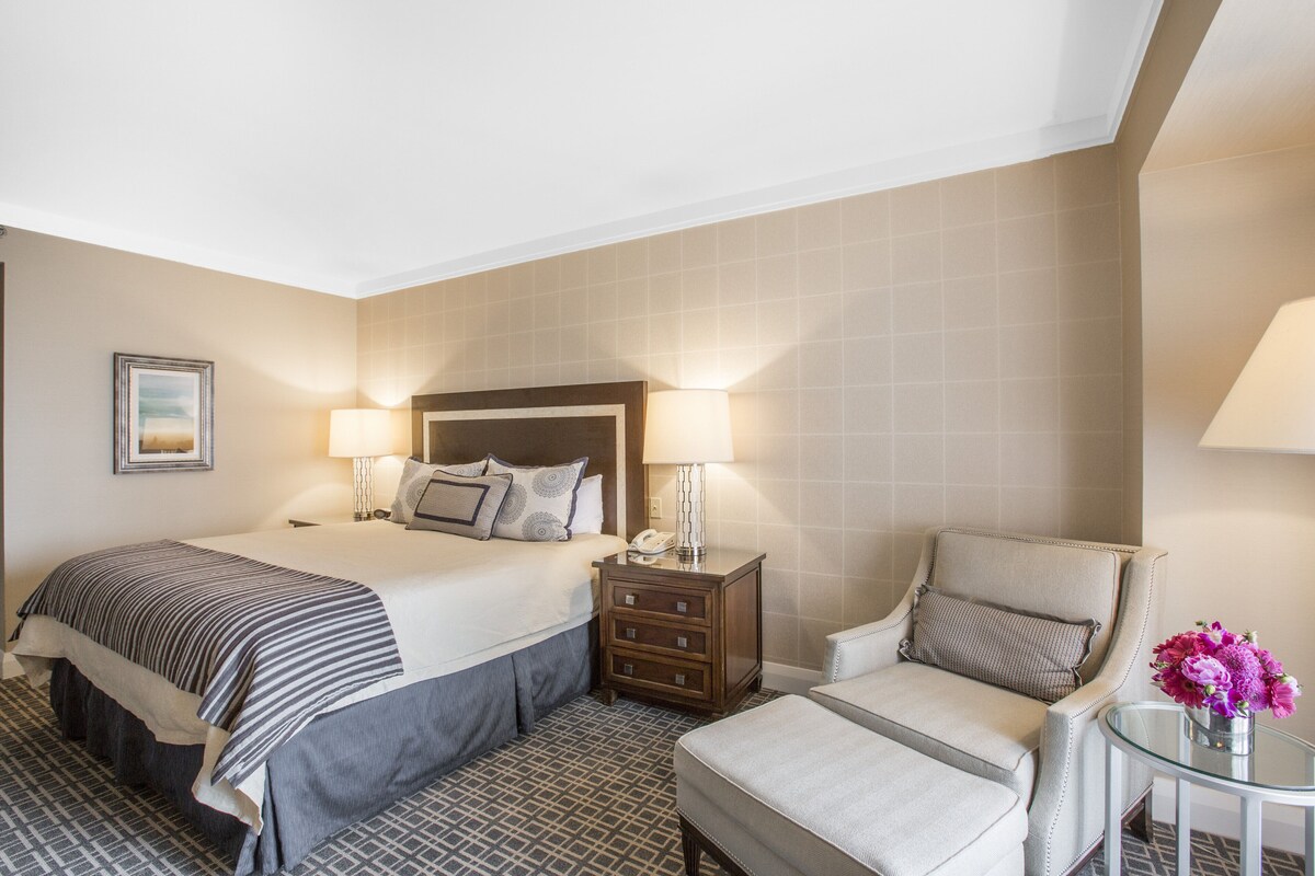 A hotel room with a large bed, a chair, a small table with a vase of flowers, and a nightstand with a lamp.
