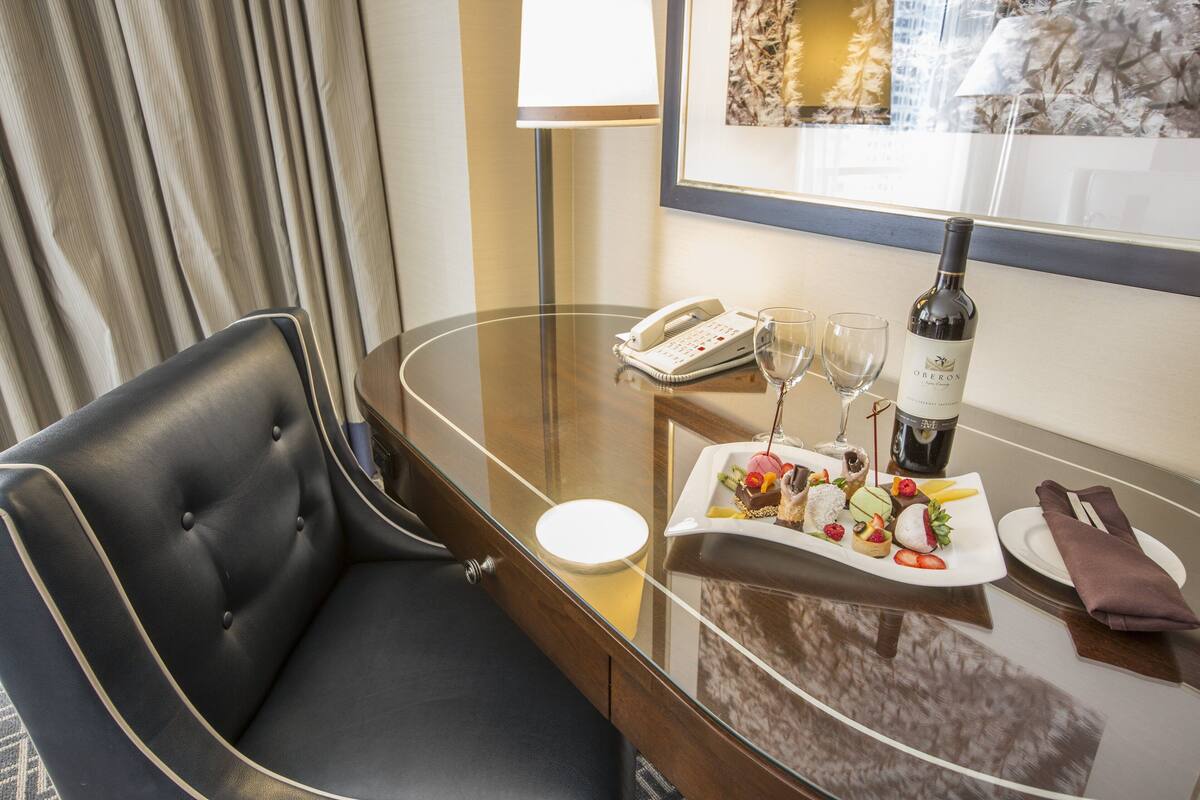 A hotel room with a glass table, a black leather chair, a telephone, wine glasses, a bottle of wine, and a plate of food.