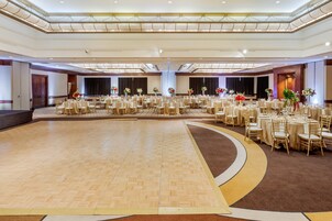 A large ballroom with a polished wooden floor, round tables with chairs, and floral centerpieces.
