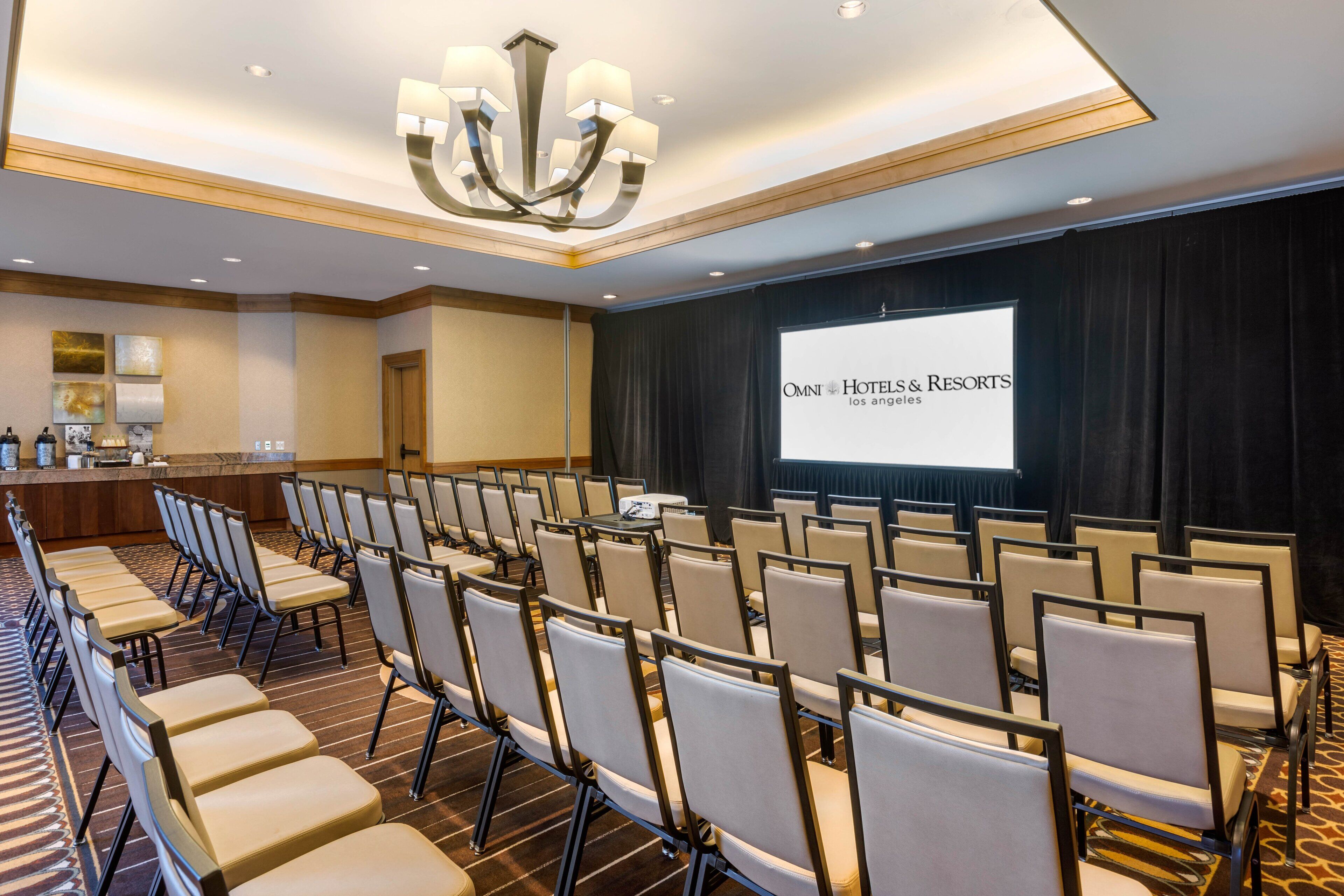Ein Konferenzraum mit Stuhlreihen, einer Projektionsleinwand, die "OMNI HOTELS & RESORTS los angeles" zeigt, und einer Kristallleuchter.