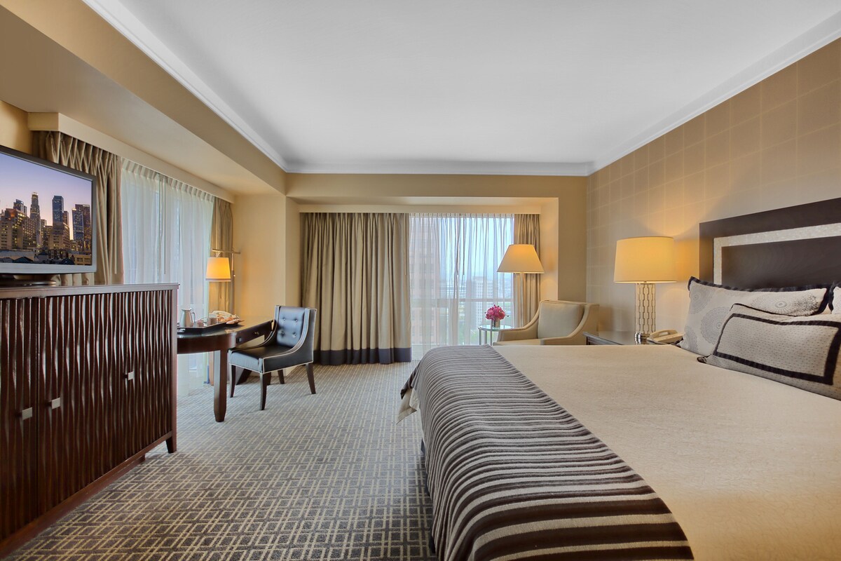 A hotel room with a large bed, a desk with a chair, a television, and a view of the city through the window.