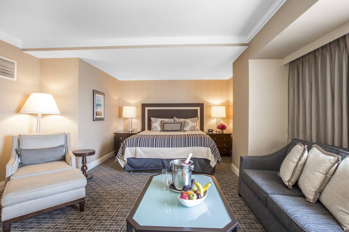 A hotel room with a bed, two chairs, a small table with a fruit bowl and a drink, and a nightstand with lamps.