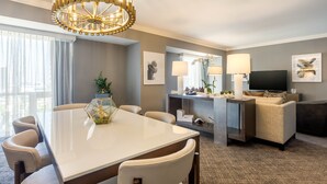 Una habitación de hotel moderna con zona de comedor, sofá, televisión y un gran ventanal con cortinas.