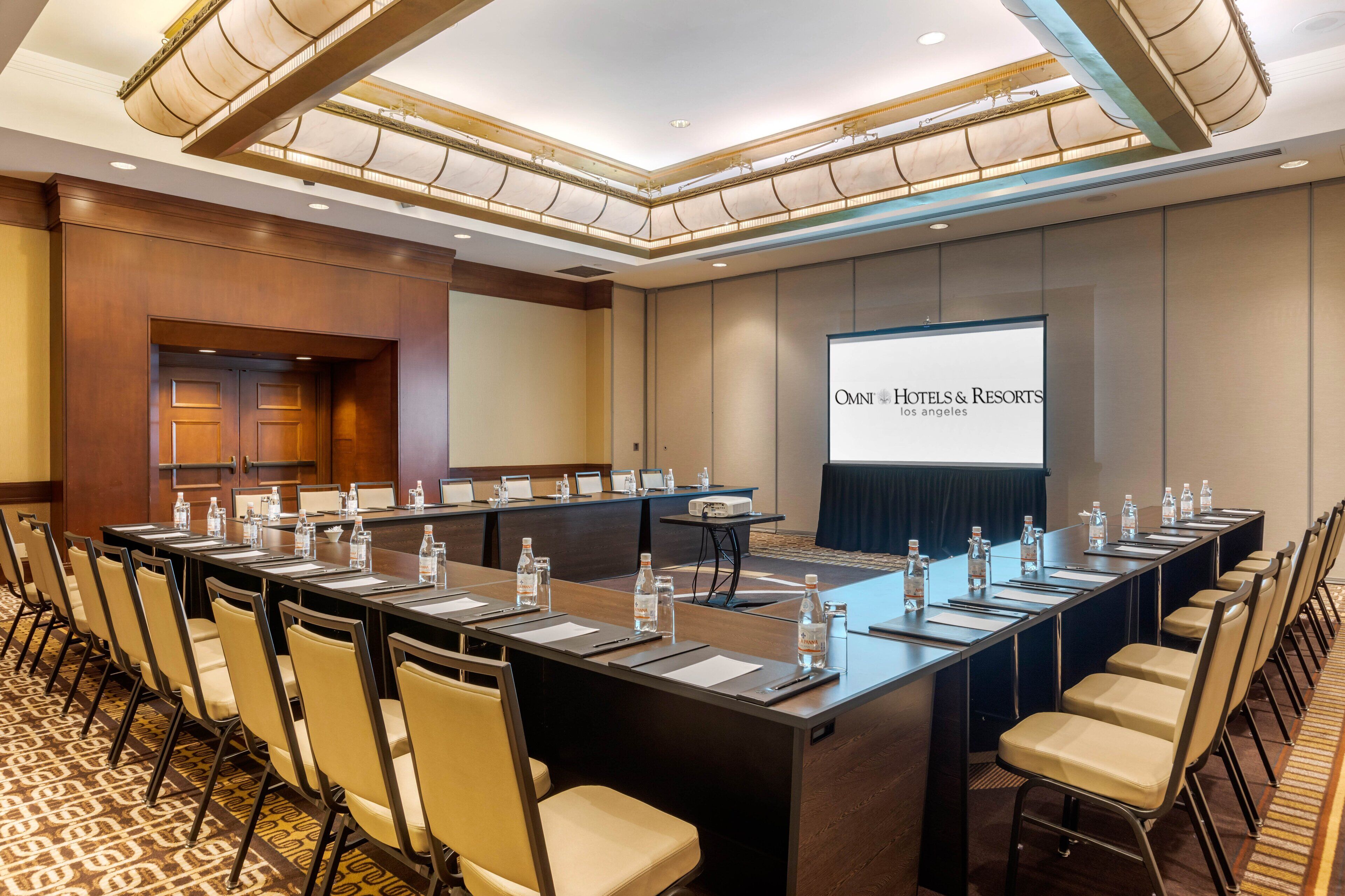 Ein Konferenzraum mit einem langen Tisch, Stühlen und einer Projektionsleinwand, die "OMNT Hotels & Resorts Los Angeles" zeigt.