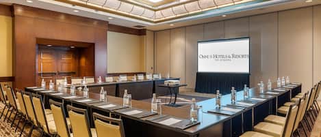 Ein Konferenzraum mit einem langen Tisch, Stühlen und einer Projektionsleinwand, die "OMNT Hotels & Resorts Los Angeles" zeigt.