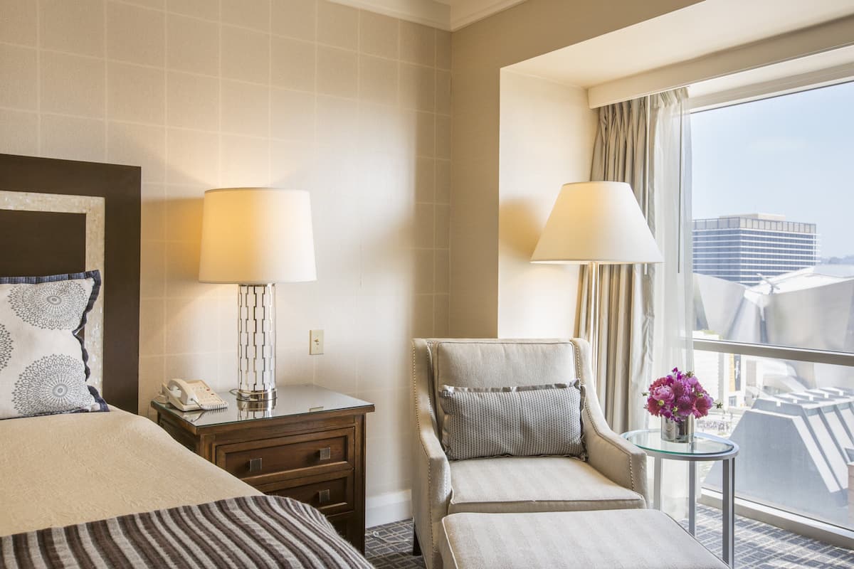 A hotel room with a bed, a desk with a lamp, a chair, a small table with flowers, and a large window with a city view.
