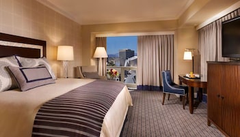 Una camera d'albergo con un letto grande, due lampade ai comodini, una scrivania con una sedia, una televisione e vista sulla città dalla finestra.
