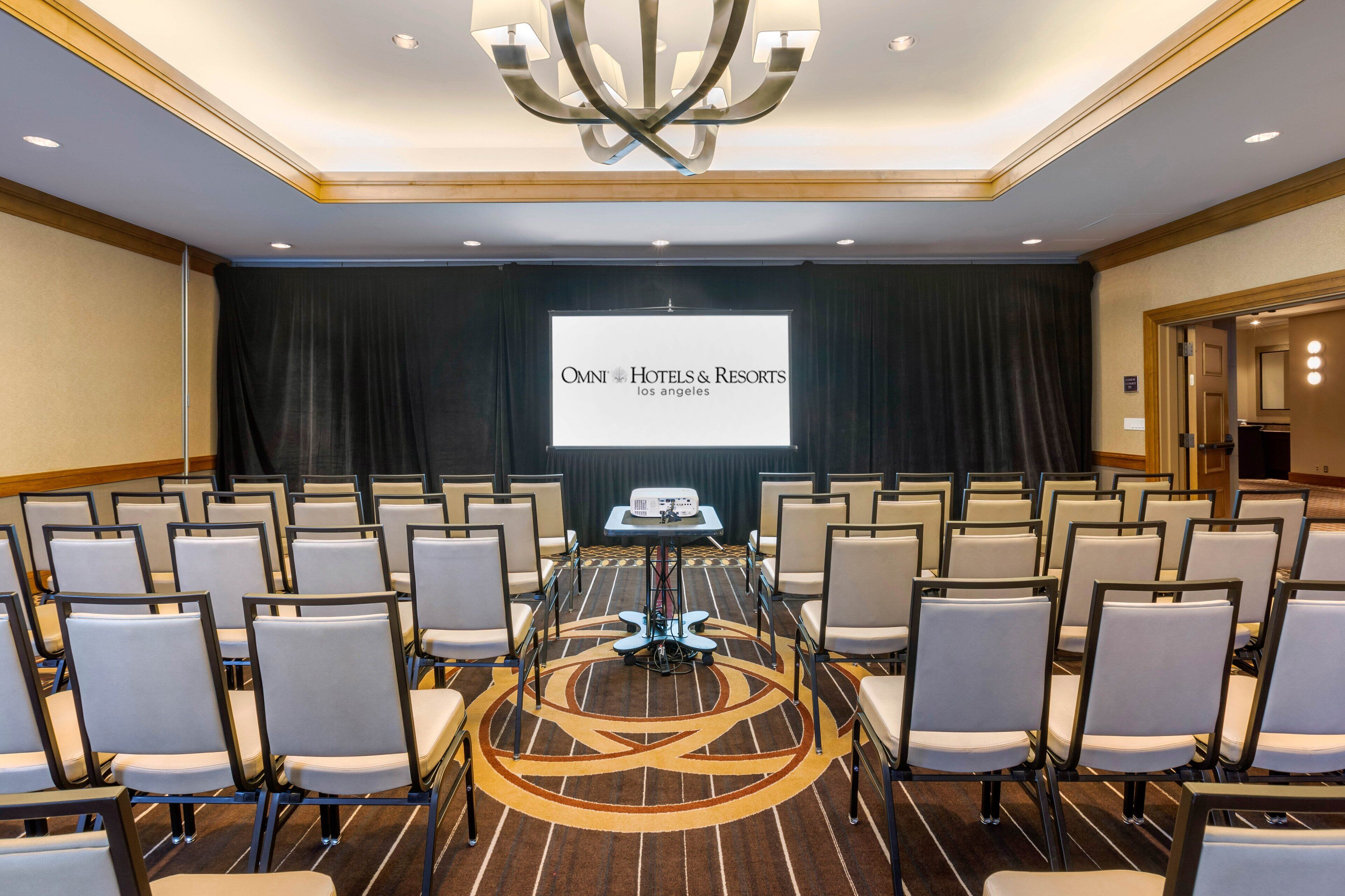 Ein Konferenzraum mit Reihen von Stühlen, einer Projektionsleinwand, auf der 'OMNI HOTELS & RESORTS Los Angeles' zu sehen ist, und einem Tisch in der Mitte mit einem Laptop.