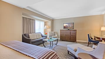 Quarto de hotel com cama, sofá, poltrona, mesinha, armário e televisão.