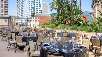 An outdoor dining area with round tables set for a meal, surrounded by chairs, with a cityscape in the background.
