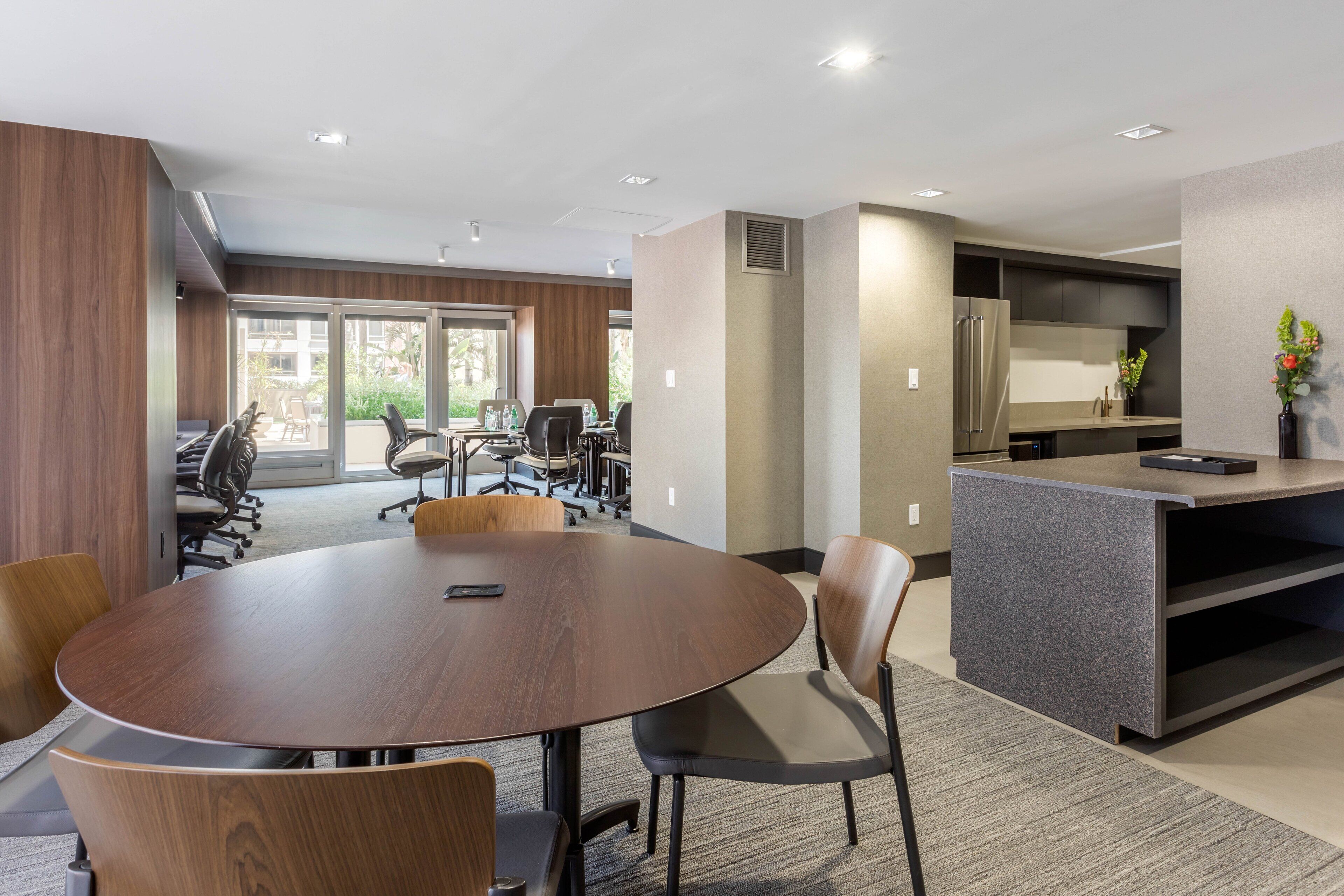 A modern conference room with a round table, chairs, and a kitchen area with a refrigerator and sink.